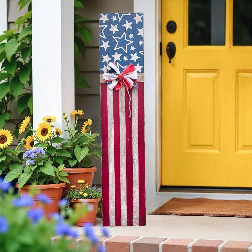 Rustic Americana flag porch leaner hand-painted navy star field and red white distressed stripes with patriotic rag ribbon bow leaning against white porch railing beside brown front door with red and pink flower pots