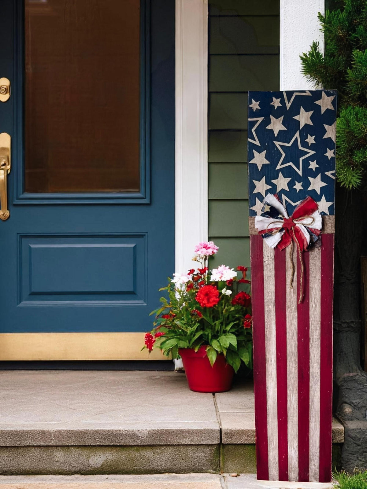 Rustic Americana flag porch leaner hand-painted navy star field and red white distressed stripes with patriotic rag ribbon bow leaning against beige siding beside brown front door with red white and blue flower pots