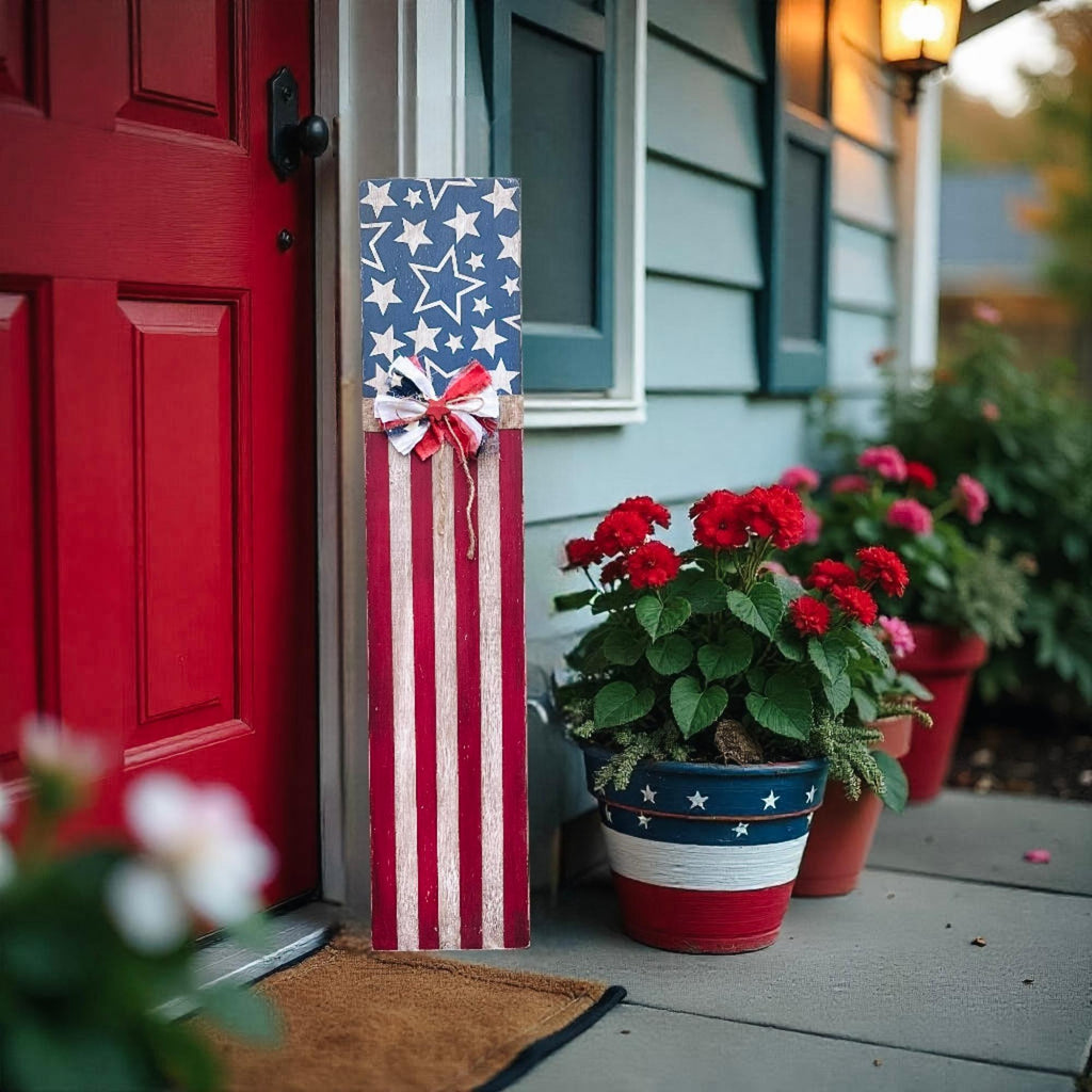Rustic Americana flag porch leaner hand-painted navy star field and red white distressed stripes with patriotic rag ribbon bow leaning against white porch column beside yellow front door with sunflowers and wildflowers