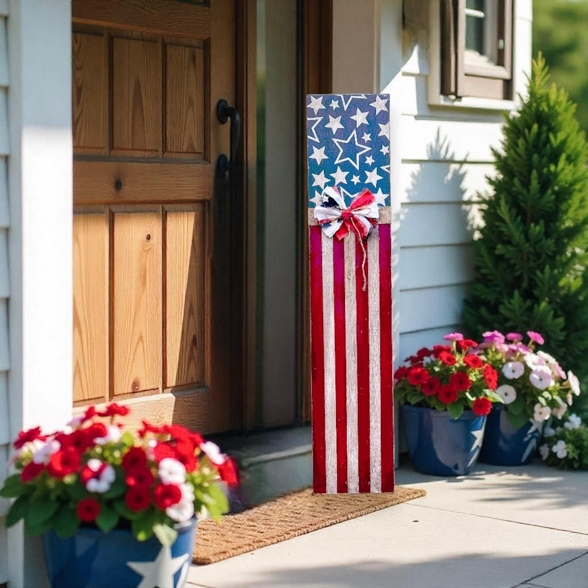 Rustic Americana flag porch leaner hand-painted navy star field and red white distressed stripes with patriotic rag ribbon bow leaning against green siding beside navy blue front door with red and white geraniums
