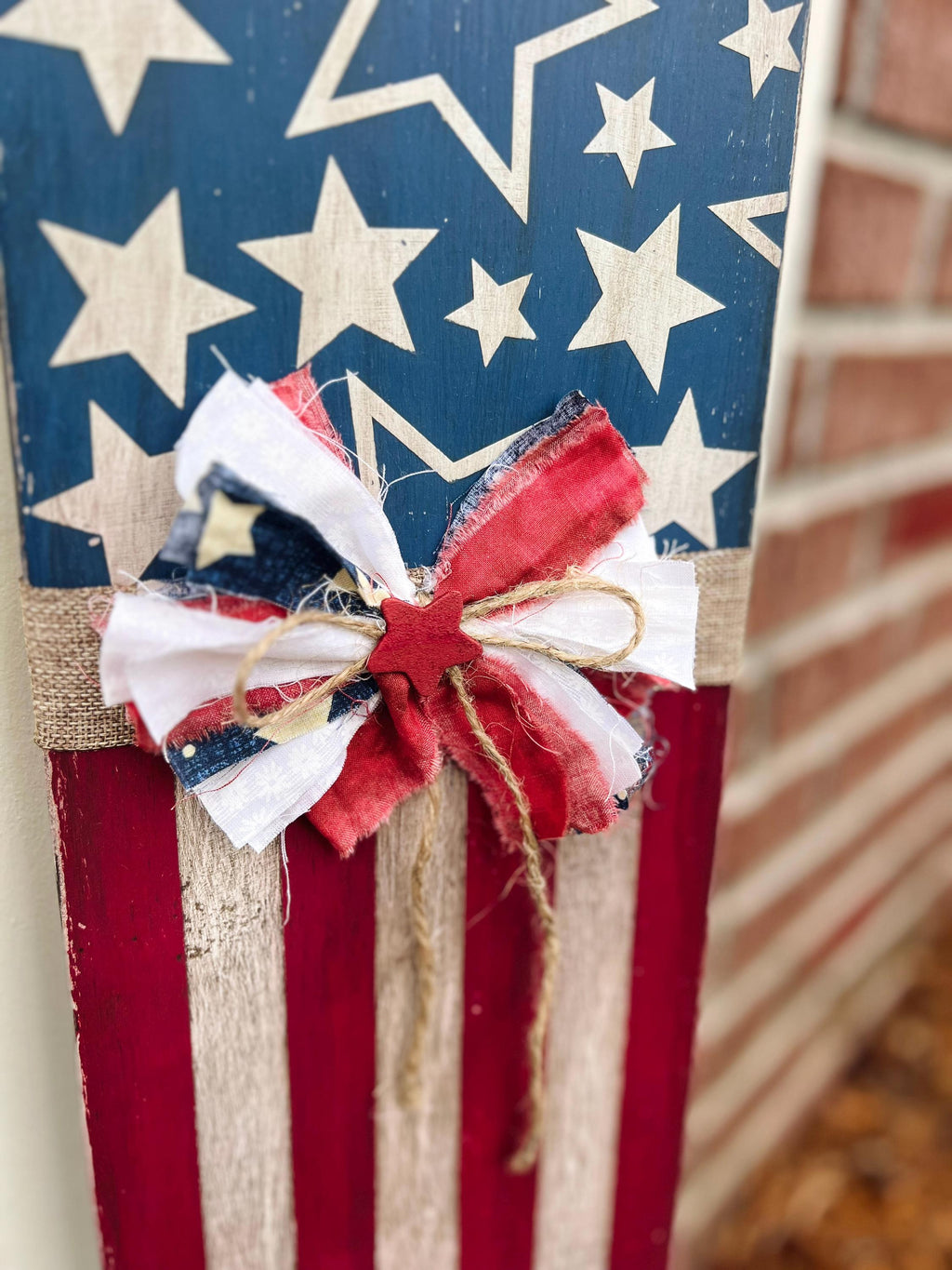 Rustic Americana flag porch leaner hand-painted navy star field and red white distressed stripes with patriotic rag ribbon bow leaning against gray siding beside red front door with red geraniums at dusk