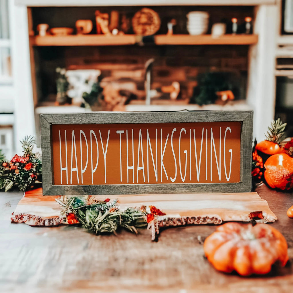 Rustic Happy Thanksgiving orange wood sign with grey frame on white wood surface with candles pine cones and fall leaves by candlelight
