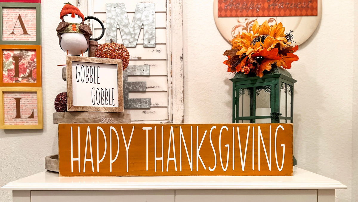 Happy Thanksgiving golden amber farmhouse wood sign with white hand-painted lettering free-standing on white console table with Gobble Gobble reversible block sign and fall floral lantern angled view