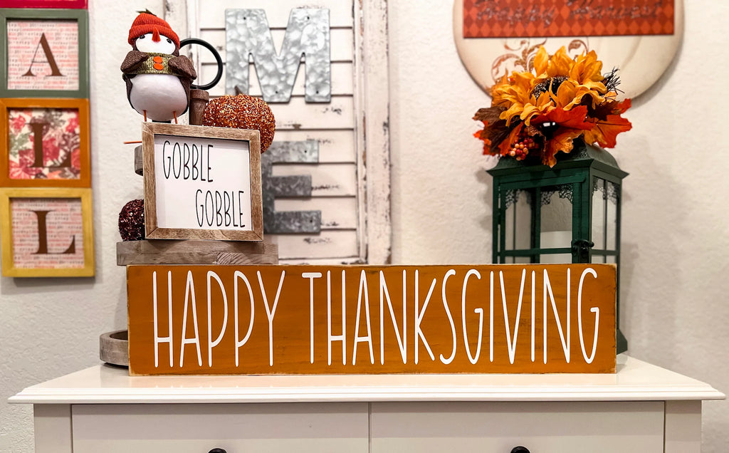 Happy Thanksgiving golden amber farmhouse wood sign with white hand-painted lettering free-standing on white console table with fall floral lantern and orange pumpkin wide shot