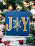 JOY navy blue Christmas wood block sign with gold serif J and Y and gold snowflake O on rustic wood surface with red and white ornaments and lit Christmas tree bokeh hero shot