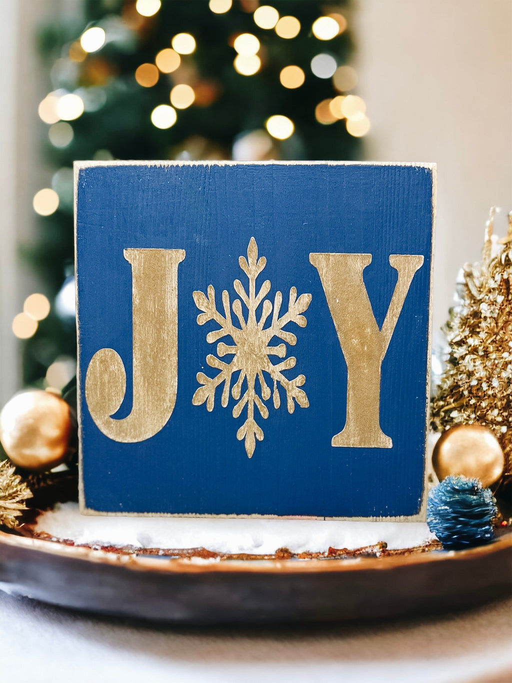 JOY navy blue Christmas wood block sign with gold serif J and Y and gold snowflake O on gold cake stand with gold glitter ornaments and lit Christmas tree bokeh front view