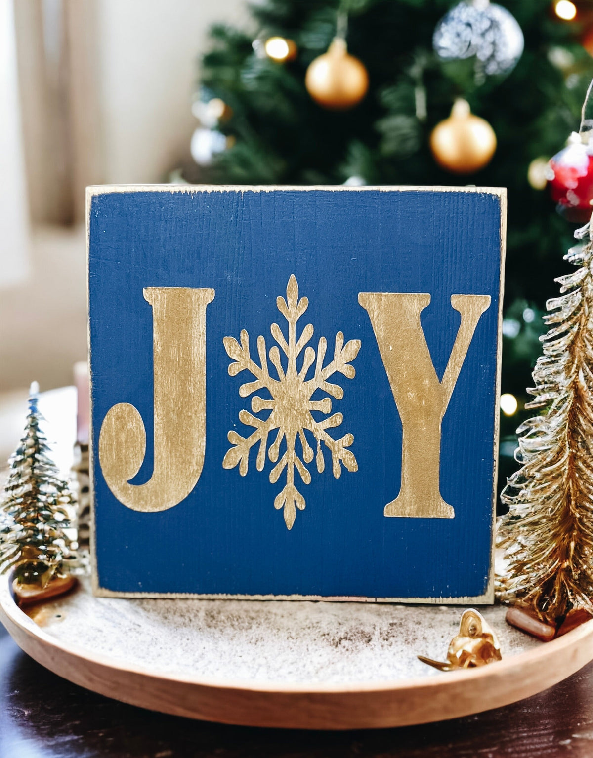 JOY navy blue Christmas wood block sign with gold serif J and Y and gold snowflake O on gold cake stand with gold glitter ornaments and lit Christmas tree bokeh side angle