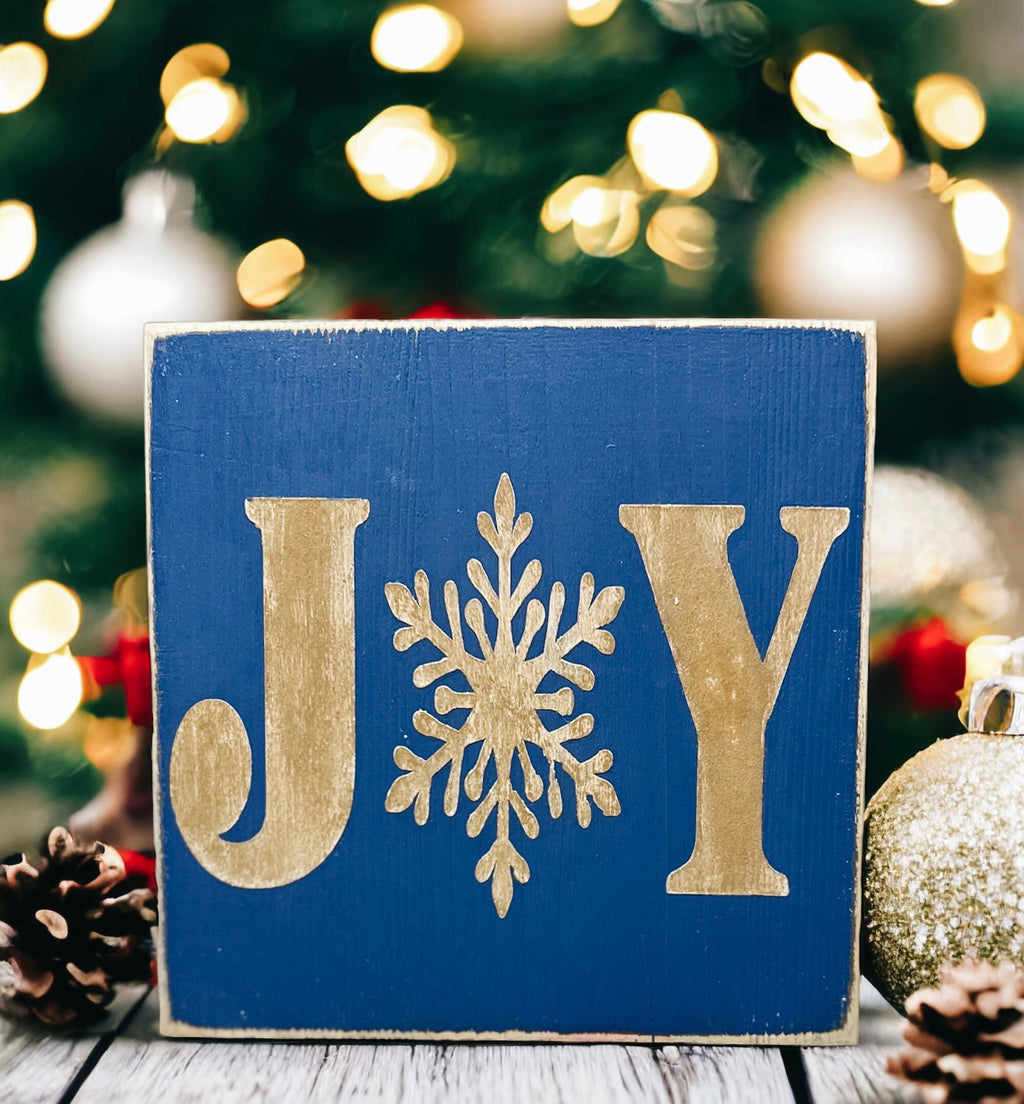 JOY navy blue Christmas wood block sign with gold serif J and Y and gold snowflake O on gold cake stand with gold glitter ornaments and white linen bokeh angled view