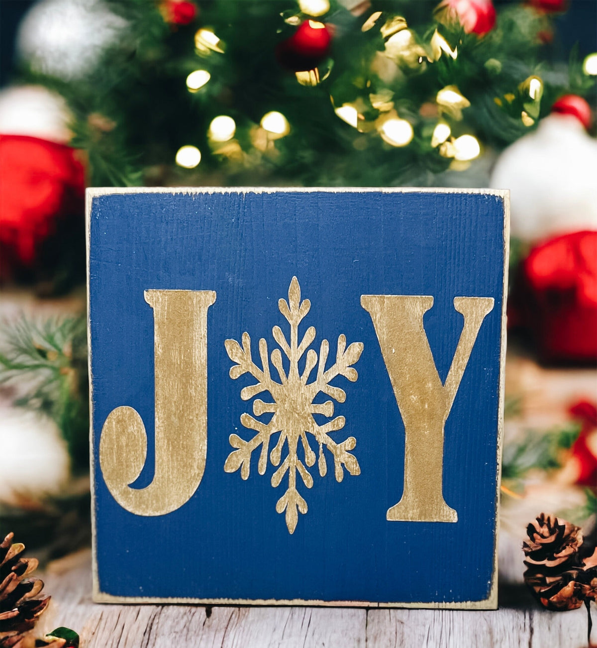 JOY navy blue Christmas wood block sign with gold serif J and Y and gold snowflake O on gold cake stand with gold glitter ornaments and lit Christmas tree bokeh