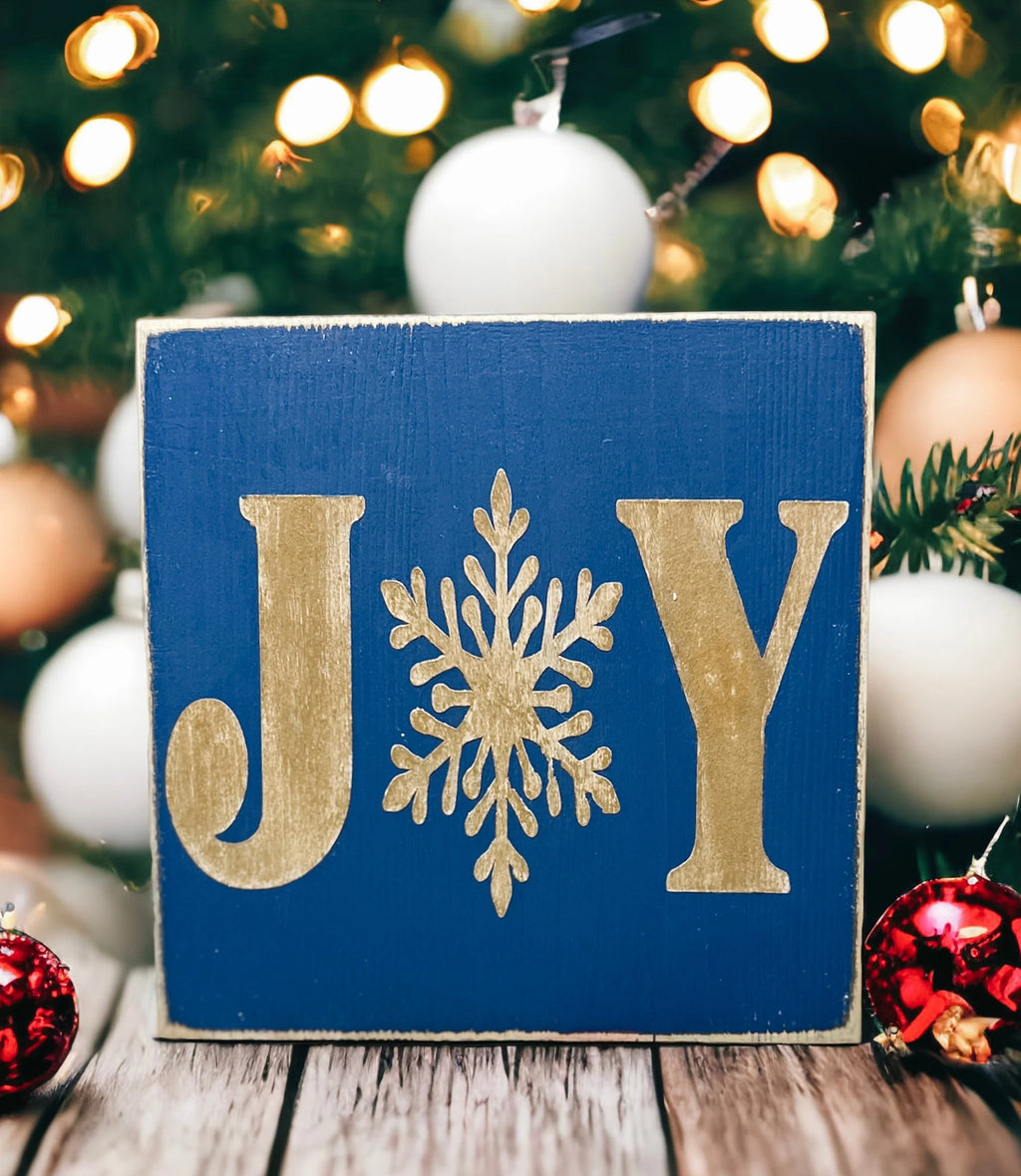 JOY navy blue Christmas wood block sign with gold serif J and Y and gold snowflake O on round wood tray with gold and teal bottle brush trees and gold ornaments bokeh close up