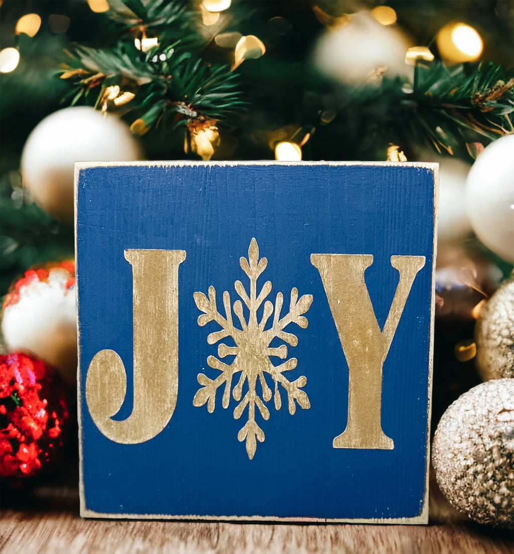 JOY navy blue Christmas wood block sign with gold serif J and Y and gold snowflake O on round wood tray with gold and silver bottle brush trees and lit Christmas tree bokeh