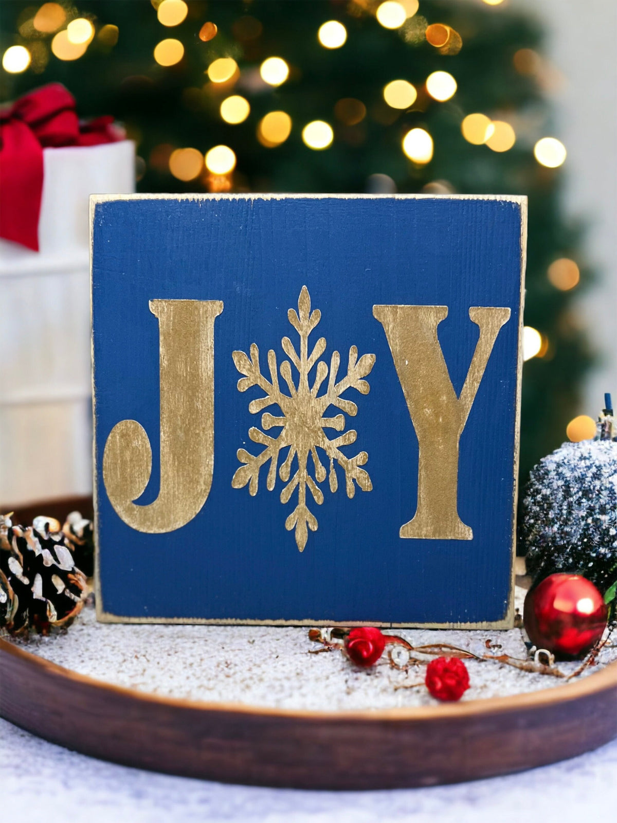 JOY navy blue Christmas wood block sign with gold serif J and Y and gold snowflake O on rustic wood surface with pine cones and gold glitter ornament bokeh wide shot