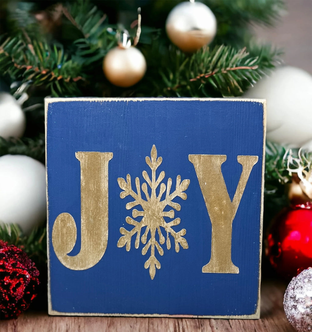 JOY navy blue Christmas wood block sign with gold serif J and Y and gold snowflake O on rustic wood surface with pine cones and lit Christmas tree with red and white ornaments bokeh