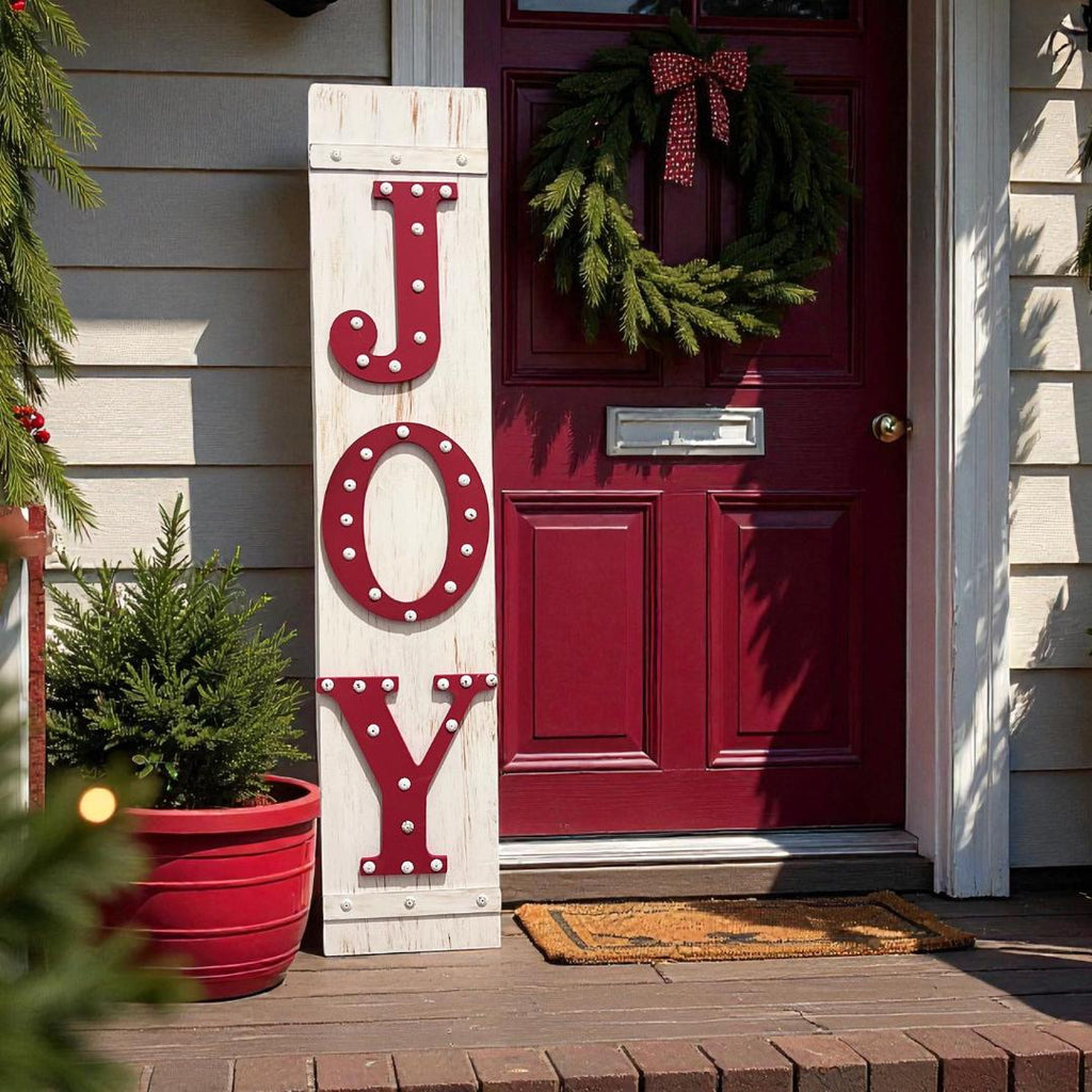 JOY porch leaner sign with red marquee letters leaning against wood front door with Christmas tree and wreath at dusk