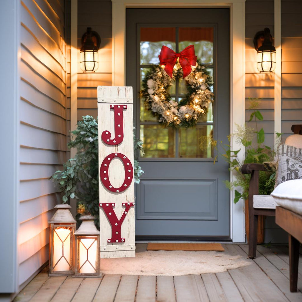 JOY porch leaner sign with red marquee letters on distressed white wood leaning against red front door with Christmas greenery and lanterns