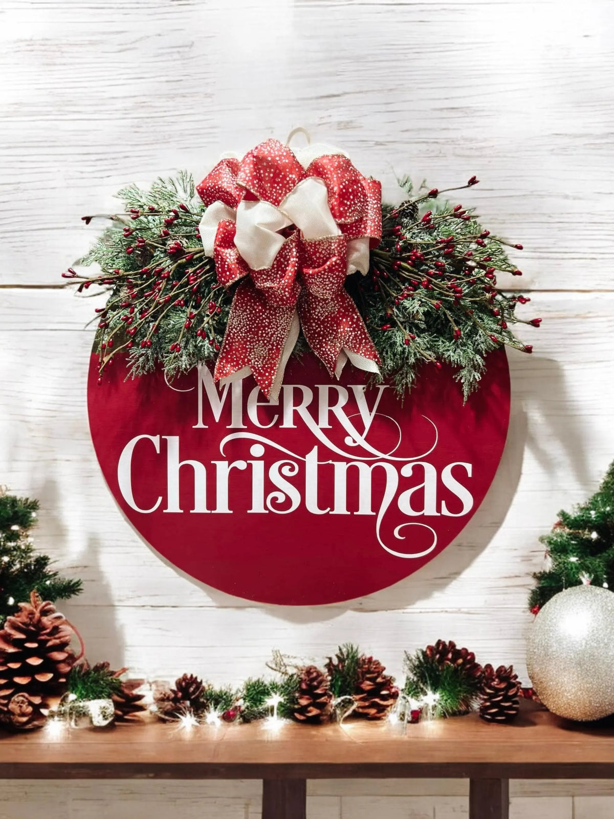 Red Merry Christmas round pine wood door hanger with frosted berry swag and red cream gold glitter bow hung on white shiplap wall above mantel with pine cone garland and string lights