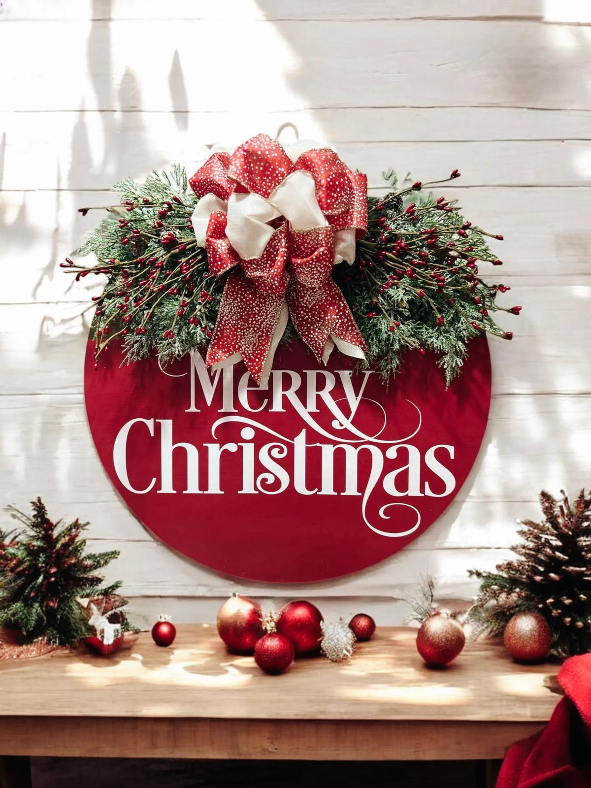 Red Merry Christmas round pine wood door hanger with frosted berry swag and red cream gold glitter bow hung on white shiplap wall above mantel with red ornaments and pine cones