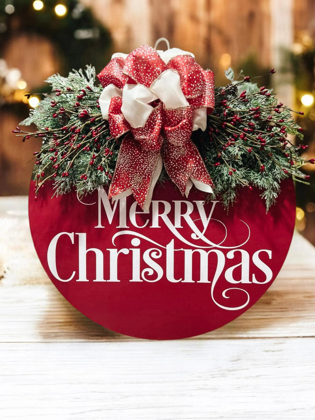 Red Merry Christmas round pine wood door hanger with frosted berry swag and red cream gold glitter bow on white wood surface product hero shot