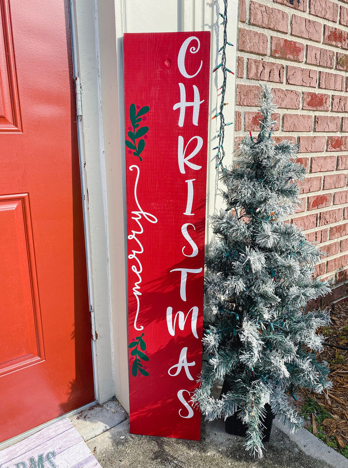 Merry Christmas red porch leaner sign with white script lettering and green holly leaning against red door with flocked Christmas tree on brick porch