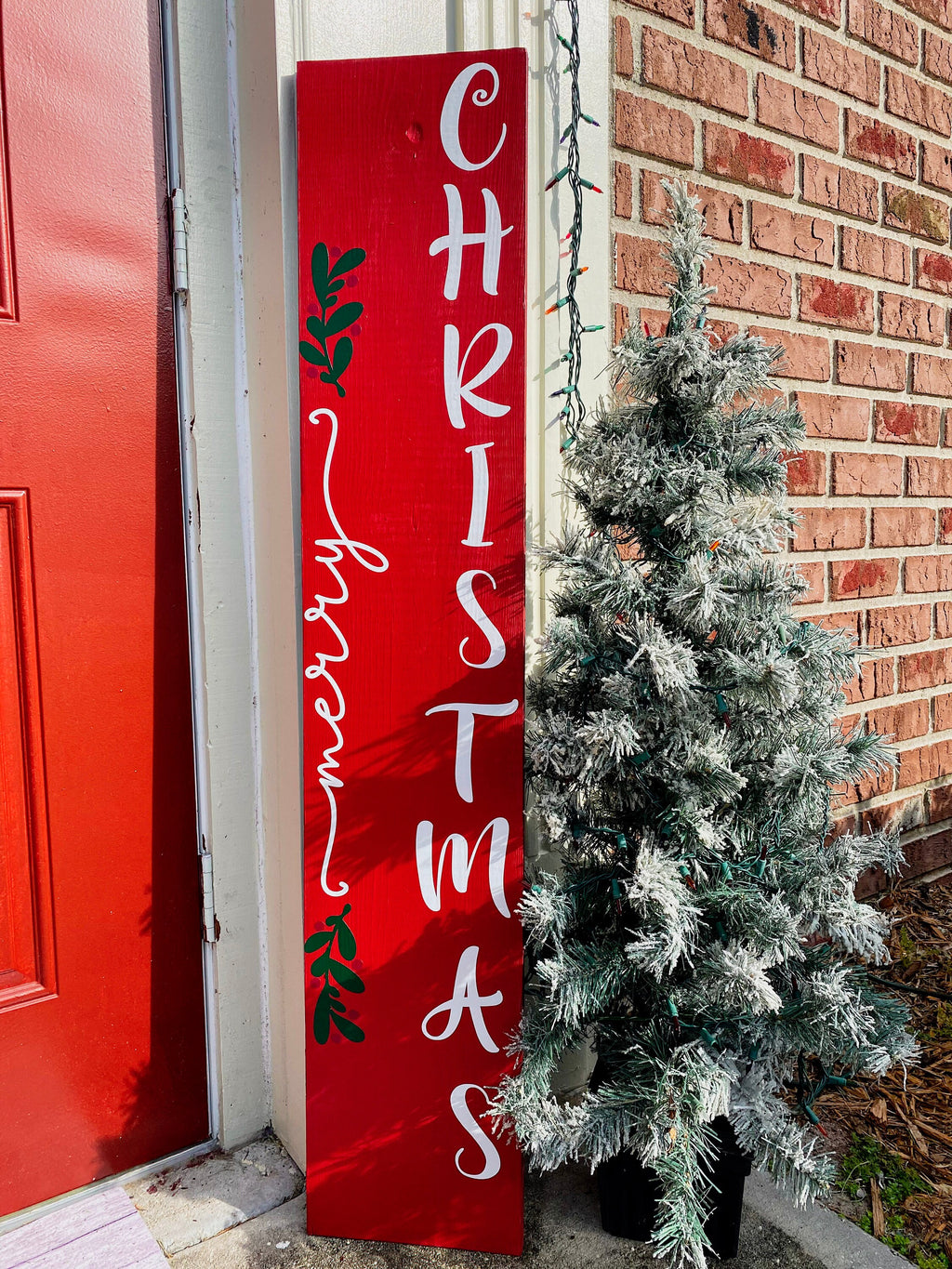 Merry Christmas red porch leaner sign with white script lettering and green holly leaning against white door frame with flocked Christmas tree on brick porch wide shot