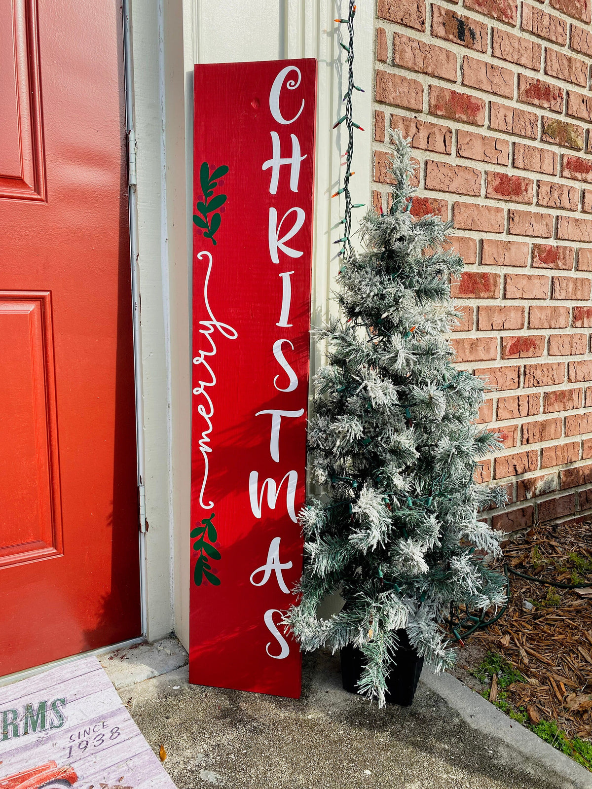 Merry Christmas red porch leaner sign with white script lettering and green holly leaning against white door frame with flocked Christmas tree on brick porch angled view