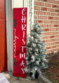 Merry Christmas red porch leaner sign with white script lettering and green holly leaning against white door frame with flocked Christmas tree on brick porch close up