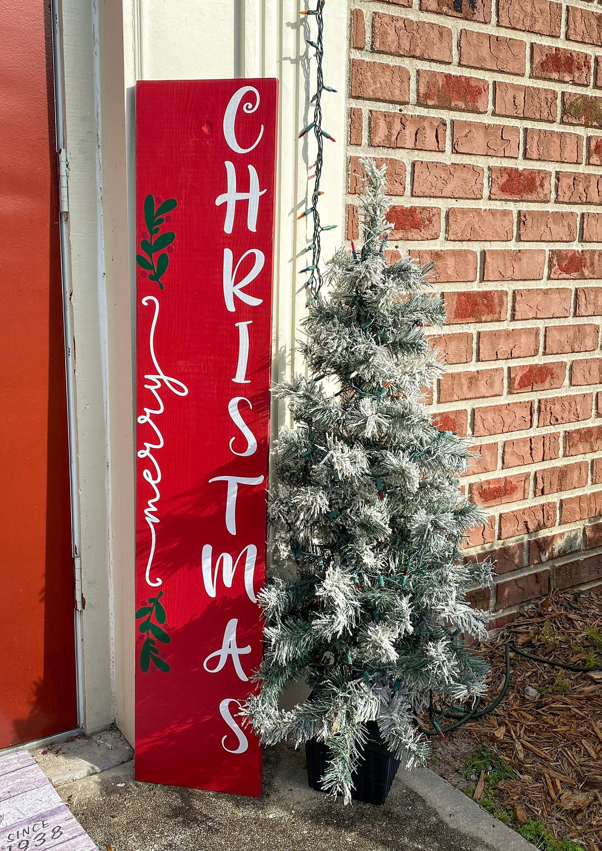 Merry Christmas red porch leaner sign with white script lettering and green holly leaning against white door frame with flocked Christmas tree on brick porch close up
