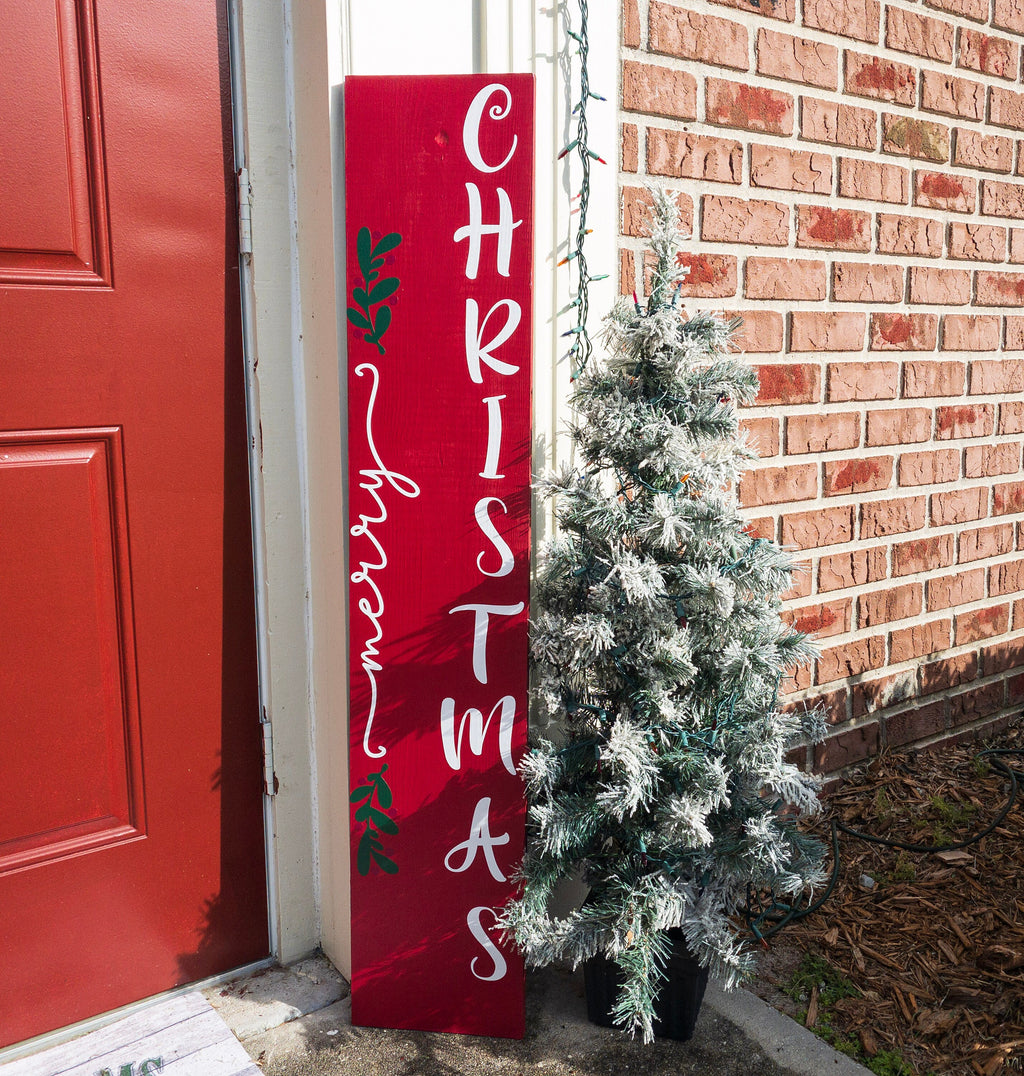 Merry Christmas red porch leaner sign with white script lettering and green holly leaning against white door frame with flocked Christmas tree on brick porch full length shot
