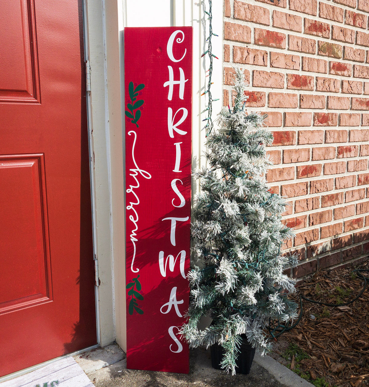 Merry Christmas red porch leaner sign with white script lettering and green holly leaning against white door frame with flocked Christmas tree on brick porch full length shot