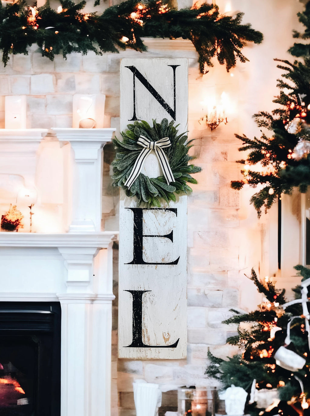Farmhouse NOEL distressed white wood sign with magnolia wreath and striped bow on white mantel with flocked bottle brush trees angled view