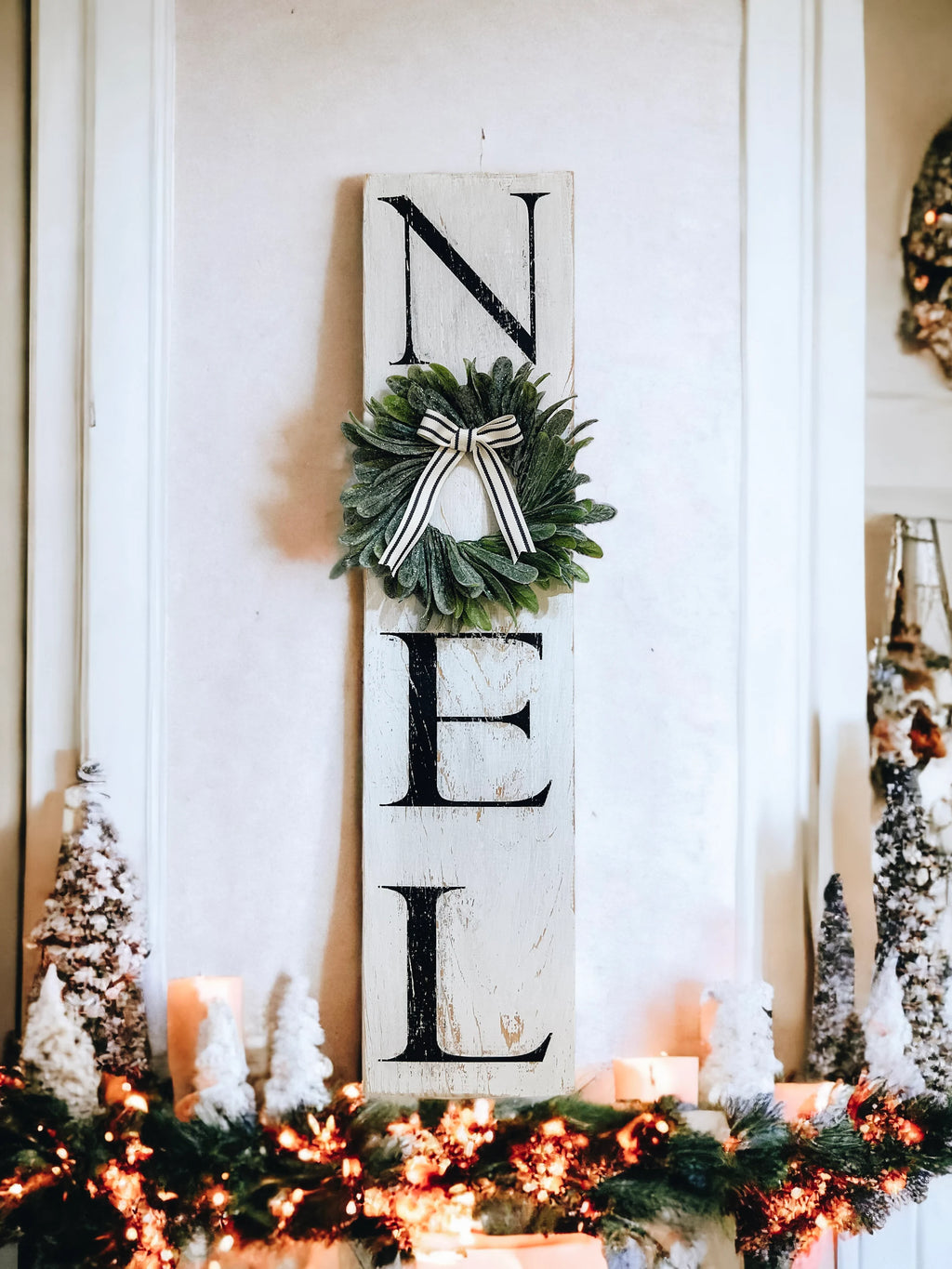 Farmhouse NOEL distressed white wood sign with magnolia wreath and striped bow on white mantel with flocked bottle brush trees close up