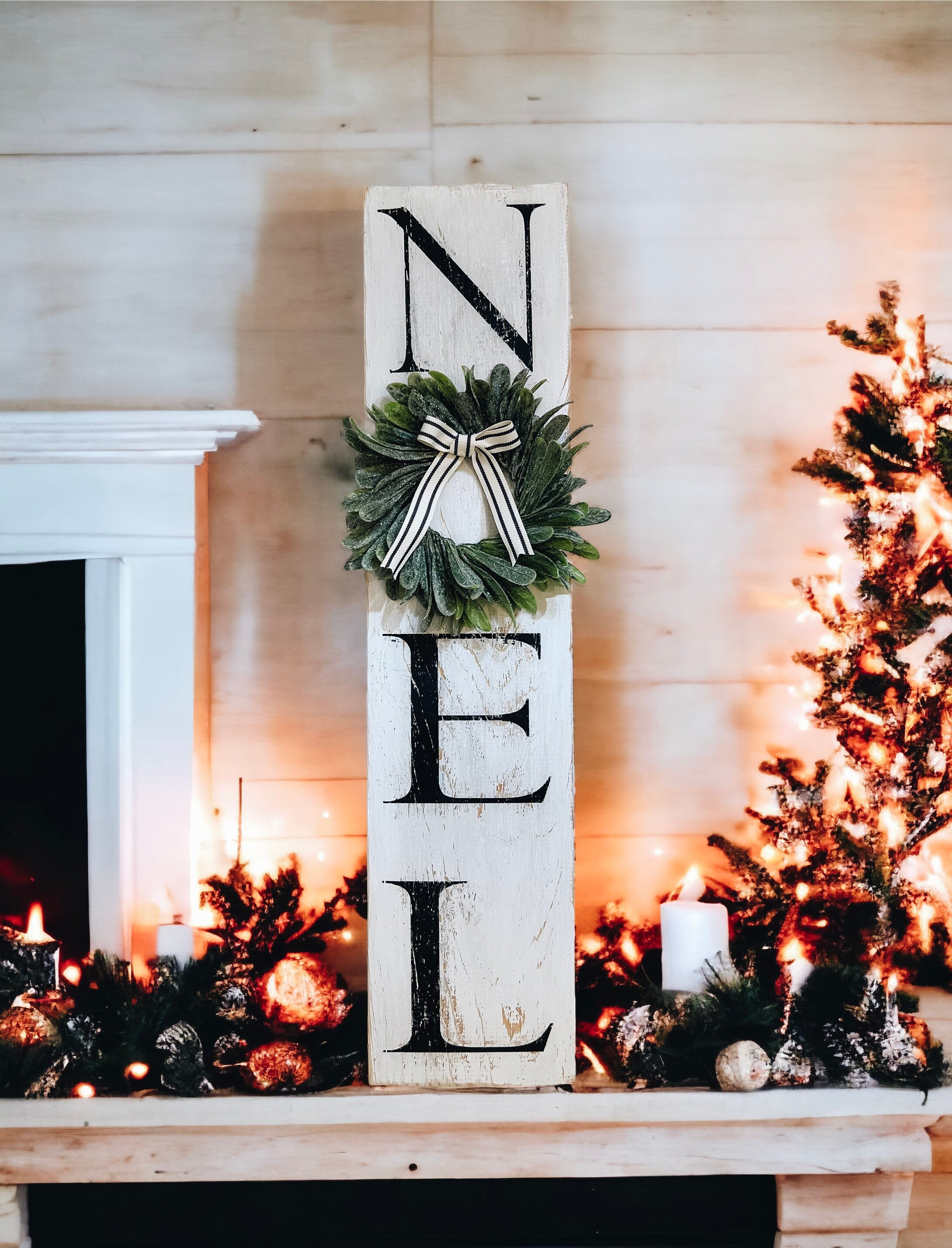 Farmhouse NOEL distressed white wood sign with magnolia wreath and striped bow on white mantel with flocked bottle brush trees and warm bokeh lights