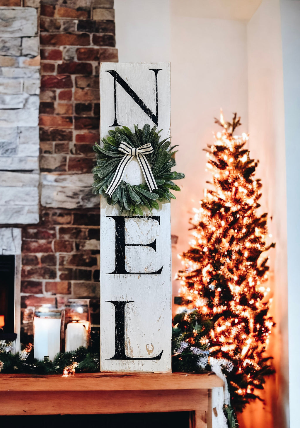 Farmhouse NOEL distressed white wood sign with magnolia wreath and striped bow on white stone fireplace mantel with lit Christmas tree and pine garland