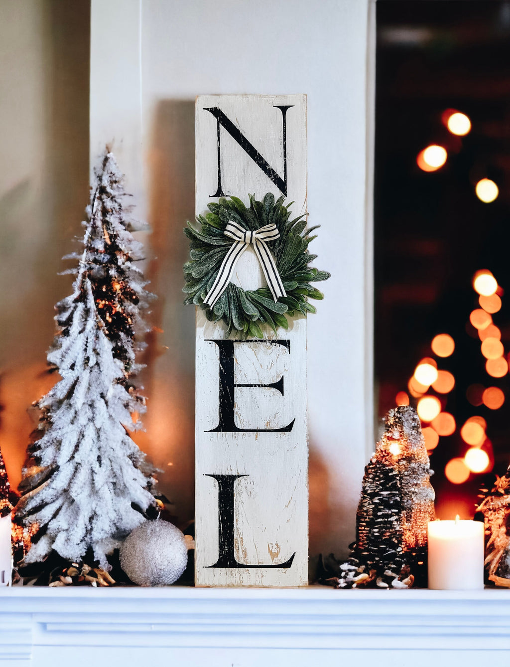 Farmhouse NOEL distressed white wood sign with magnolia wreath and striped bow hung on white door with flocked bottle brush trees and candles below