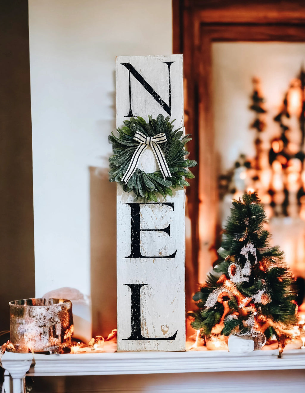 Farmhouse NOEL distressed white wood sign with magnolia wreath and striped bow on rustic wood mantel with brick fireplace and lit Christmas tree
