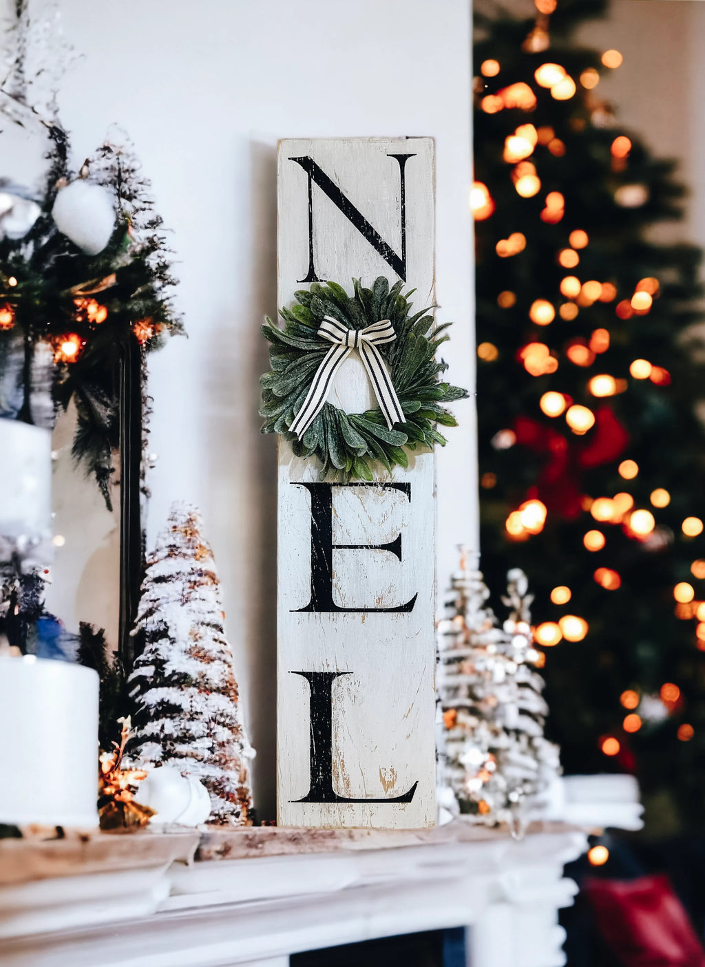 Farmhouse NOEL distressed white wood sign with magnolia wreath and striped bow on white mantel with flocked bottle brush trees wide lifestyle shot
