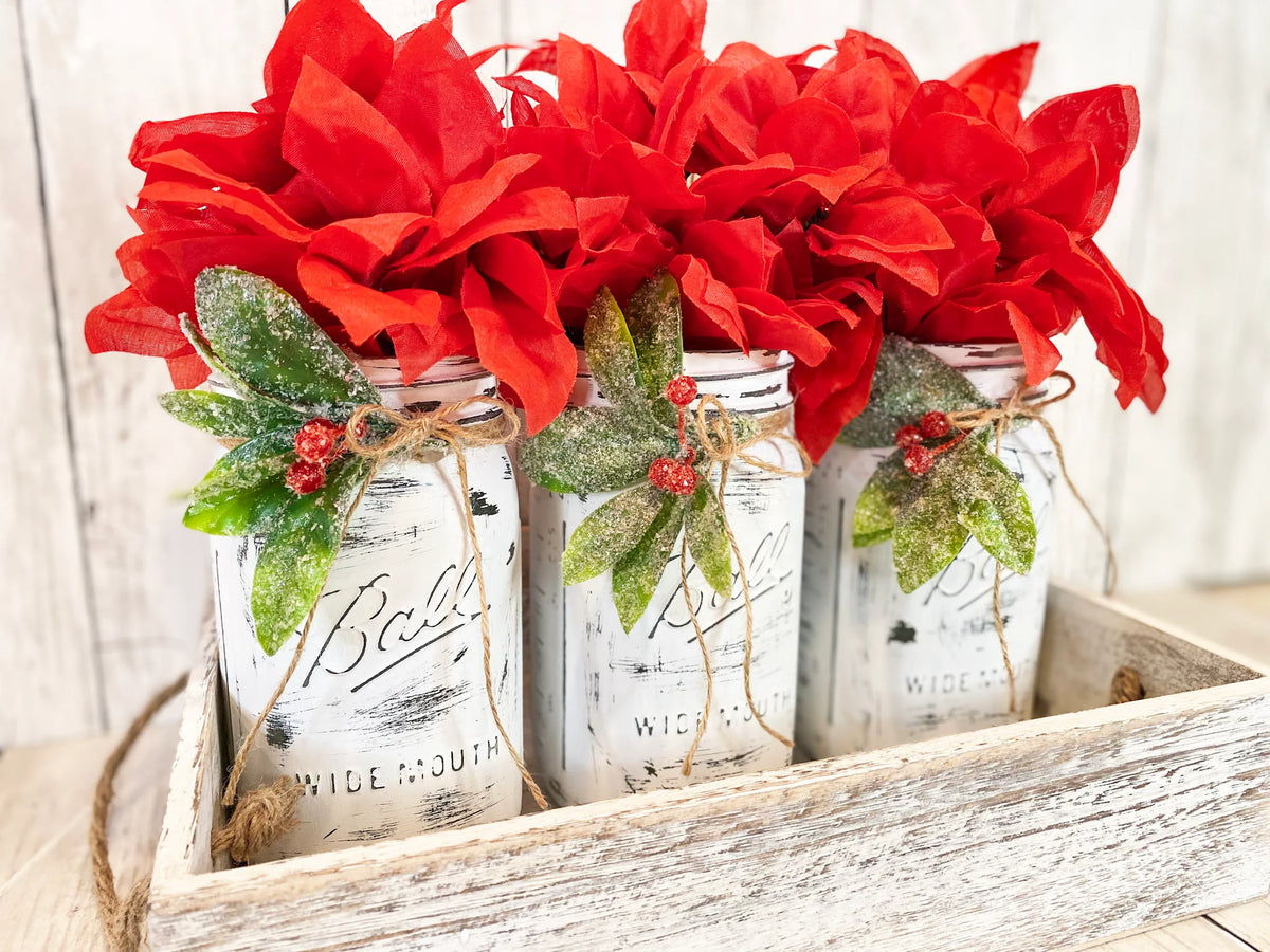 Three distressed white Ball mason jars with red poinsettia bushes holly berries and jute ties in whitewashed wood tray close up of holly and jute detail