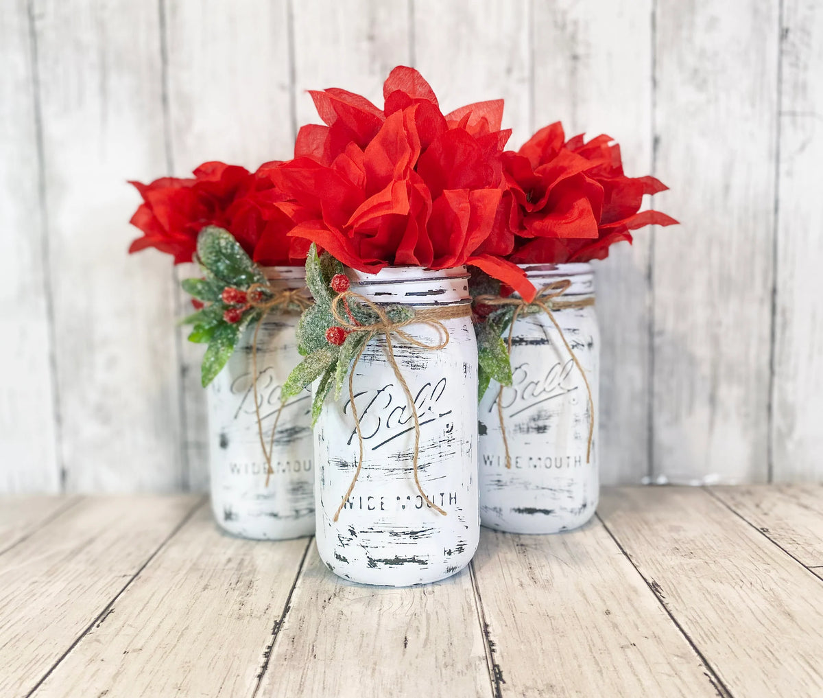 Three distressed white Ball mason jars with red poinsettia bushes holly berries and jute ties in whitewashed wood tray on white wood surface front view