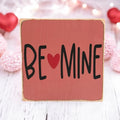 Be Mine Valentine wood block sign on rustic wood surface with red roses and bokeh lights in background