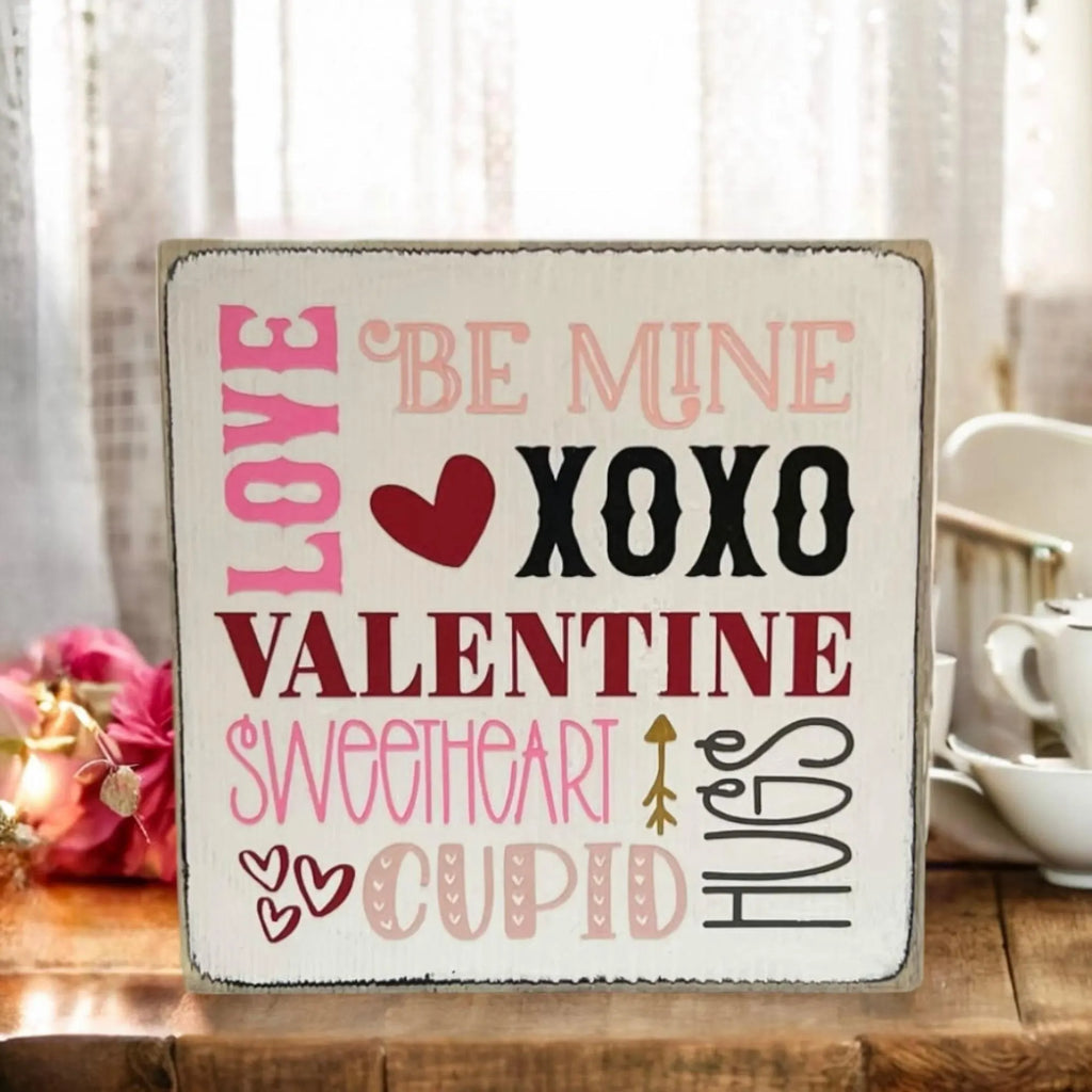 Valentine sayings wood block sign with Love Be Mine XOXO Valentine Sweetheart Cupid Hugs in pink crimson and black on white pine on rustic wood surface with pink rose petals and warm candlelight wide shot
