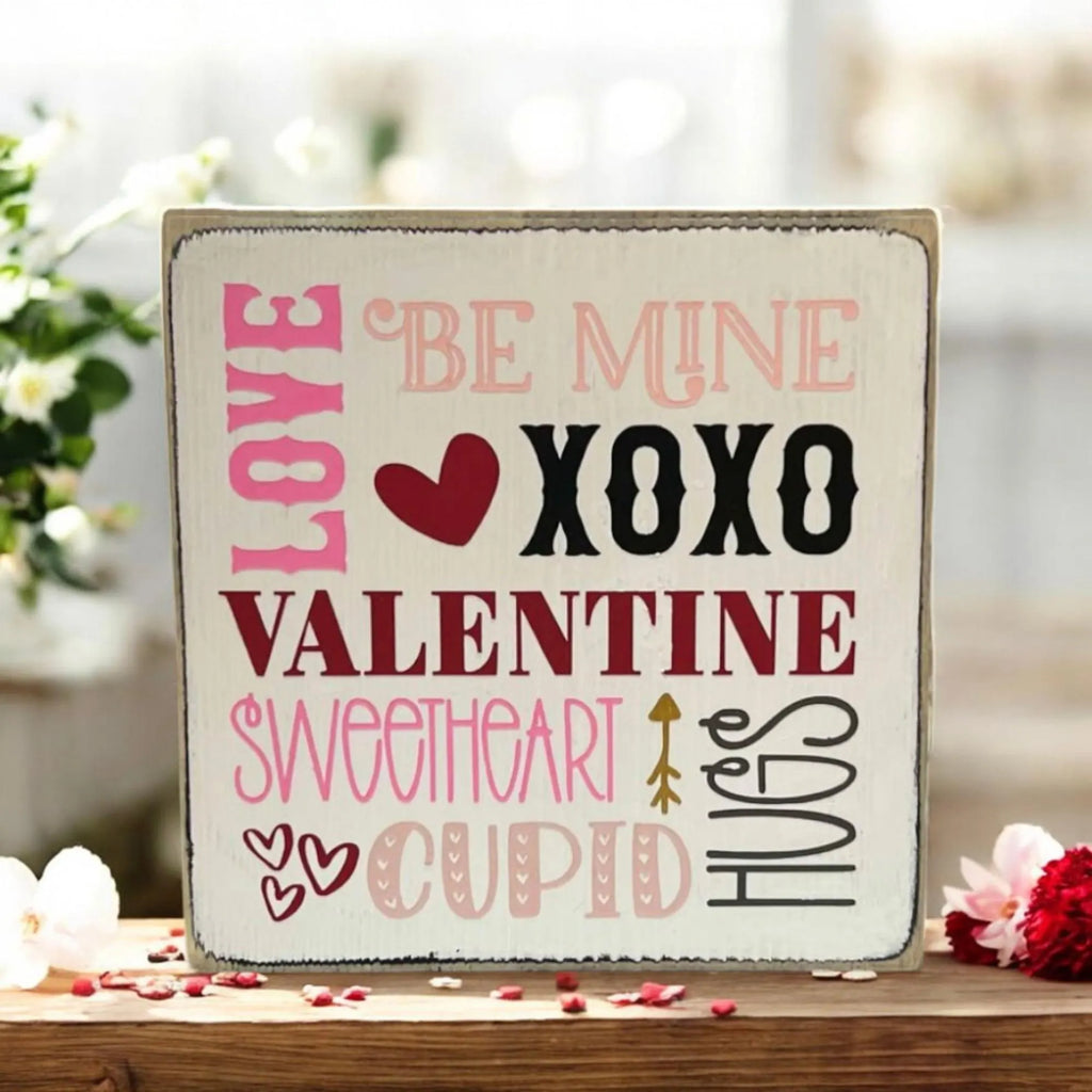 Valentine sayings wood block sign with Love Be Mine XOXO Valentine Sweetheart Cupid Hugs in pink crimson and black on white pine on rustic wood surface with pink rose petals and warm candlelight bokeh