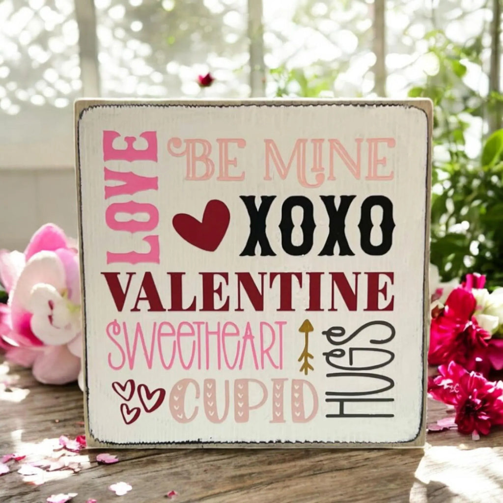 Valentine sayings wood block sign with Love Be Mine XOXO Valentine Sweetheart Cupid Hugs in pink crimson and black on white pine on rustic wood surface with pink roses and lit candles hero shot