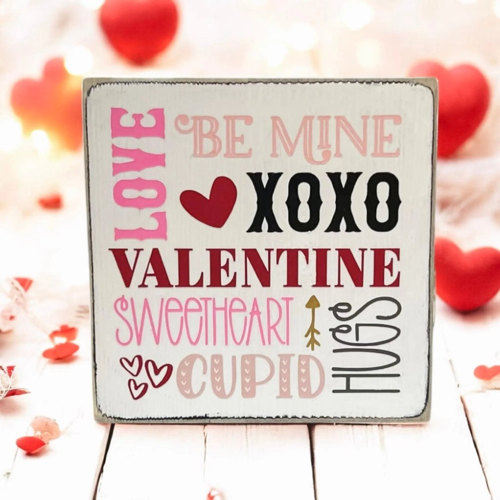 Valentine sayings wood block sign with Love Be Mine XOXO Valentine Sweetheart Cupid Hugs in pink crimson and black on white pine on white shiplap surface with white teacup and pink roses lifestyle shot