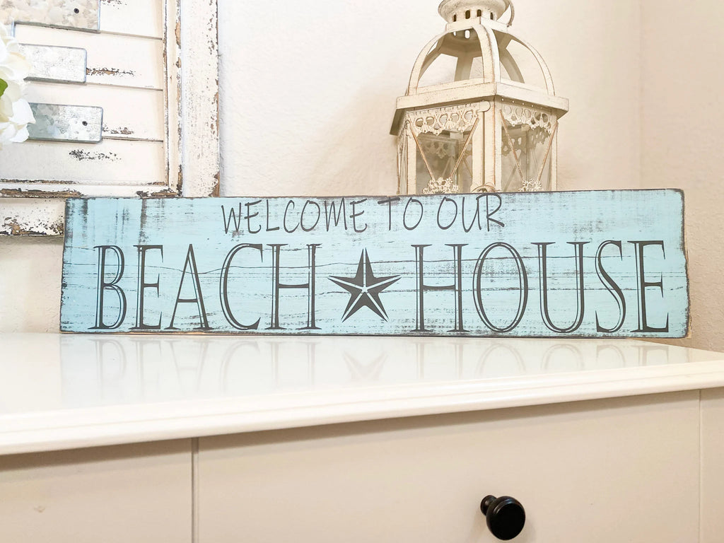 Welcome to Our Beach House reclaimed wood sign leaning against tree trunk outdoors in natural light