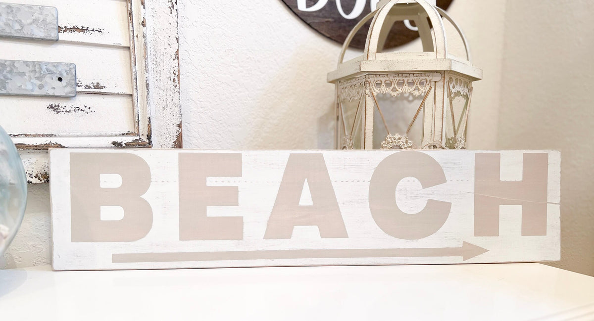Reclaimed wood Beach arrow sign in distressed white with sandy taupe lettering on white console table with vintage lantern and glass float full front view