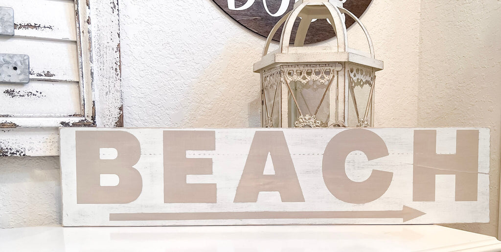 Reclaimed wood Beach arrow sign in distressed white with sandy taupe lettering on white console table with vintage lantern and glass float wide lifestyle shot
