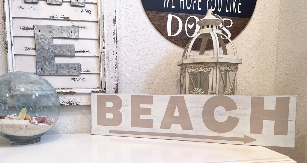 Reclaimed wood Beach arrow sign in distressed white with sandy taupe lettering on white console table with vintage lantern and glass float side angle