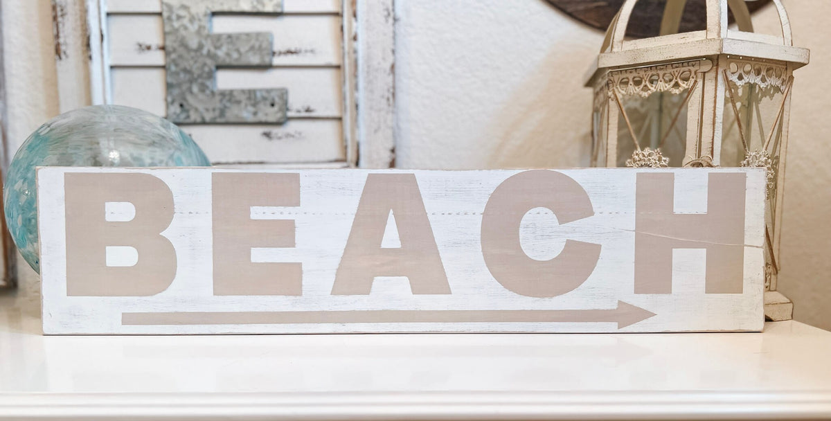 Reclaimed wood Beach arrow sign in distressed white with sandy taupe lettering on white console table with vintage lantern and glass float wide shot