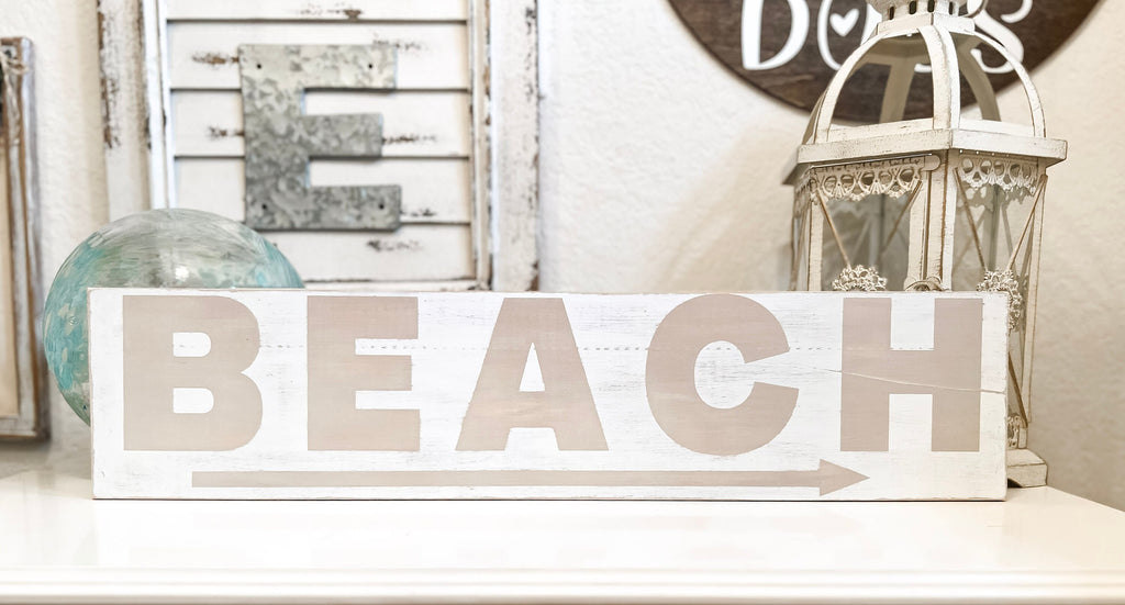 Reclaimed wood Beach arrow sign in distressed white with sandy taupe lettering on white console table with vintage lantern and glass float hero shot