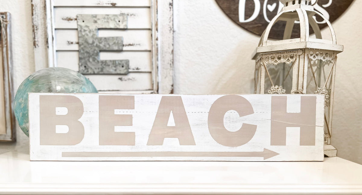 Reclaimed wood Beach arrow sign in distressed white with sandy taupe lettering on white console table with vintage lantern and glass float hero shot